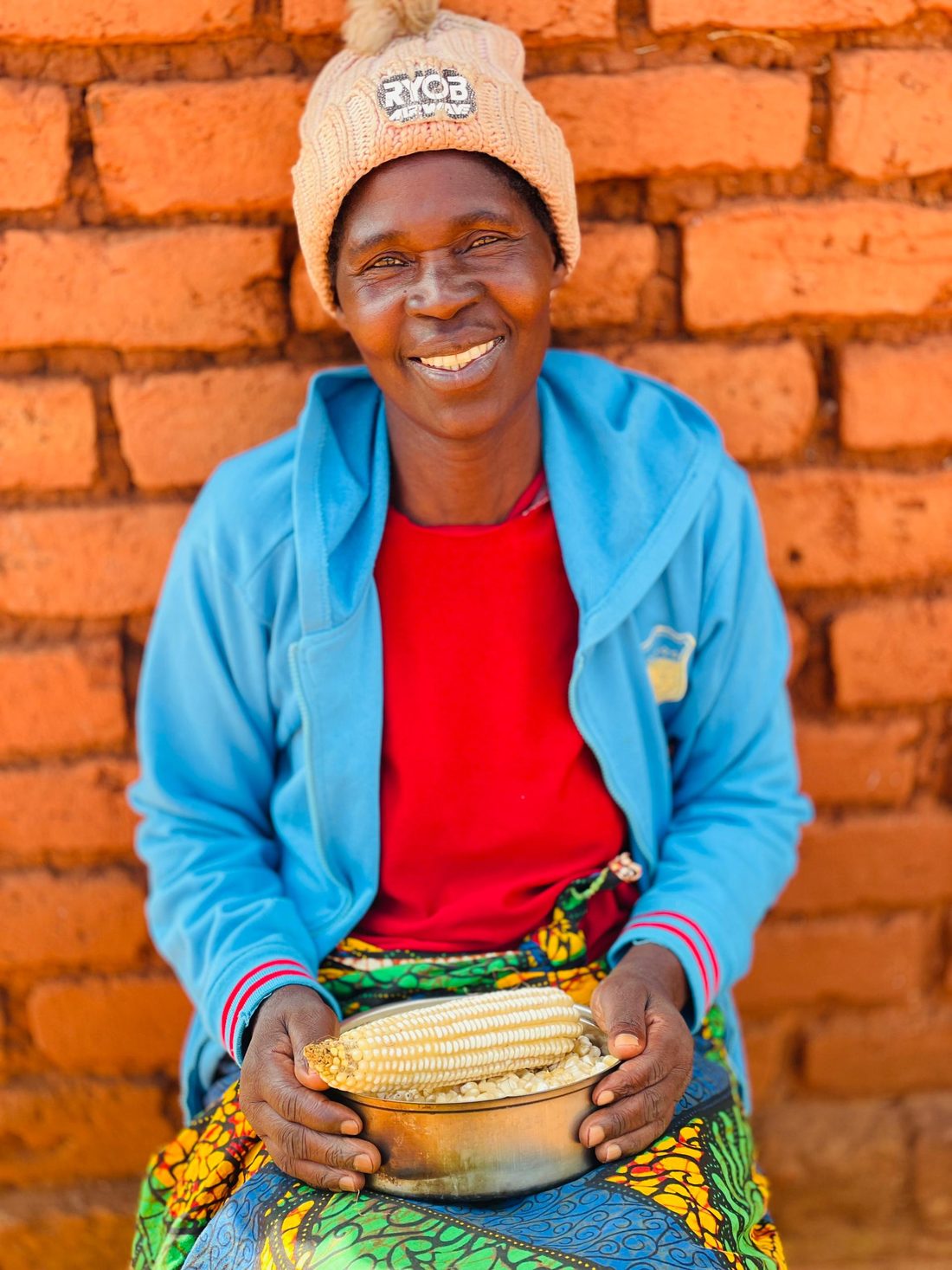 A woman sitting against a brick wall, smiling, holding a bowl of freshly harvested maize