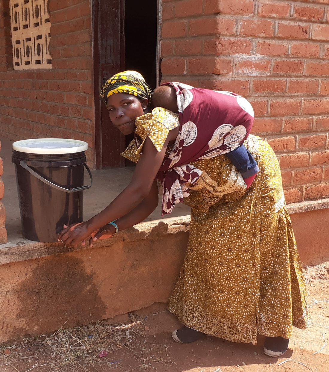 A woman washing her hands at a household water filter, her baby on her back
